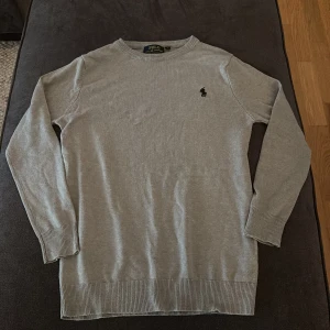 Grå tröja från Polo Ralph Lauren - Säljer en stilren grå tröja från Polo Ralph Lauren med broderad logga på bröstet. Tröjan har långa ärmar och ribbade muddar vid ärmslut och nederkant. Perfekt för en klassisk look.