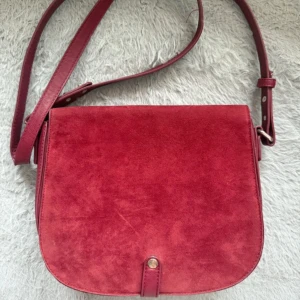 Röd axelväska från Gina Tricot - Snygg röd axelväska från Gina Tricot i mocka och skinn. Väskan har en justerbar axelrem och en praktisk dragkedja inuti för säker förvaring. Perfekt för att ge en färgklick till din outfit.