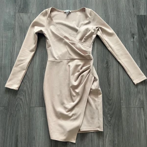 Beige omlottklänning från NLY One - Snygg beige omlottklänning från NLY One i en elegant design. Klänningen har långa ärmar och en draperad detalj på sidan som ger en fin siluett. Perfekt för en stilren look.