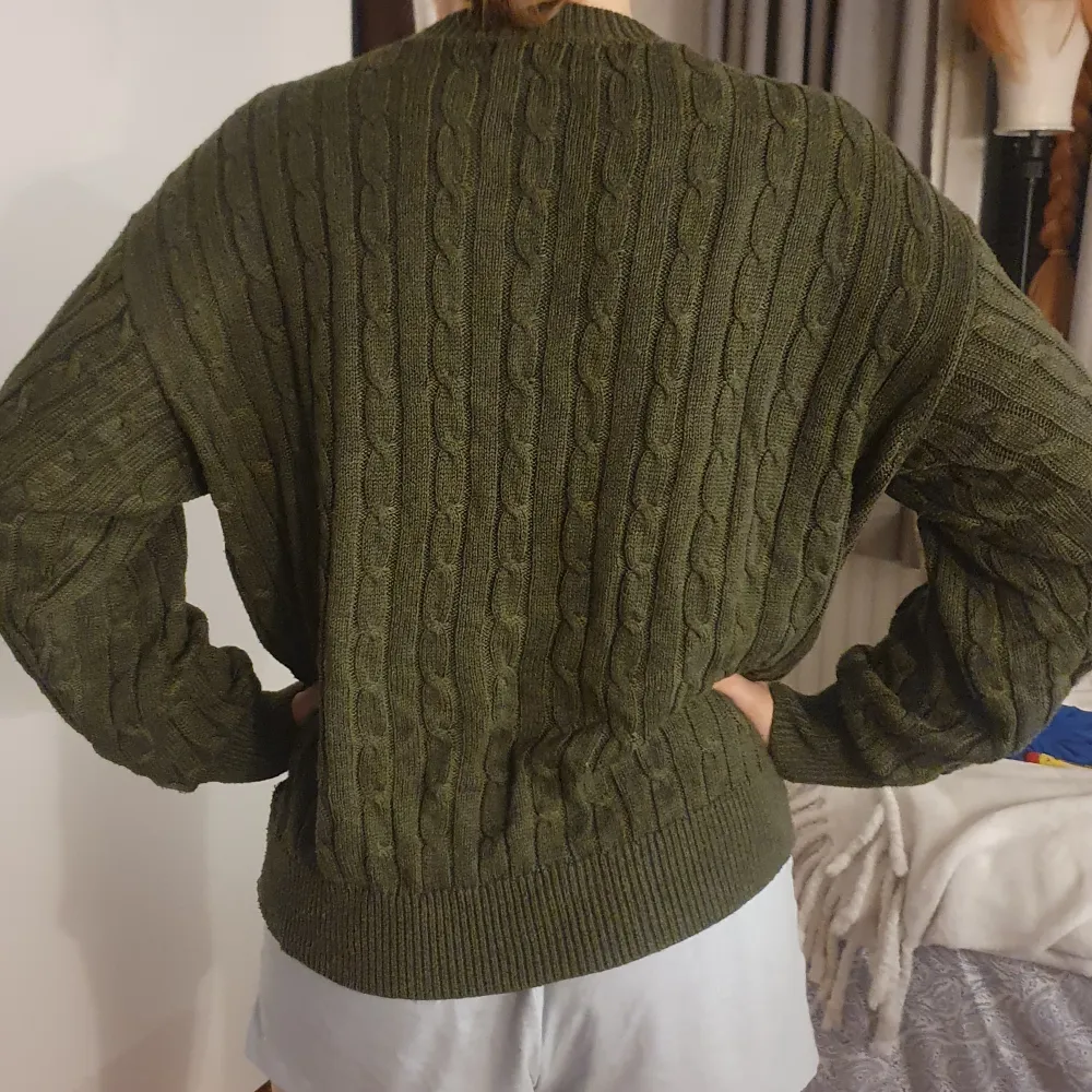 Säljer en olivgrön stickad tröja med kabelstickat mönster. Tröjan har långa ärmar och ribbade muddar vid ärmslut och nederkant. . Neuletakit & Villapaidat.
