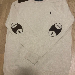 Beige tröja från Polo Ralph Lauren - Säljer en stilren beige tröja från Polo Ralph Lauren med bruna detaljer på axlarna och armbågarna. Tröjan har långa ärmar och ett diskret Polo-logotyp broderat på bröstet. Perfekt för en avslappnad och elegant look.