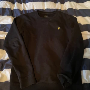 Svart tröja från Lyle & Scott - Säljer en stilren svart tröja från Lyle & Scott med ett broderat gult emblem på bröstet. Tröjan har långa ärmar och en klassisk rund halsringning. Perfekt för en avslappnad stil.
