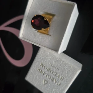 Caroline svedbom classic drop ring  - Elegant guldfärgad ring från Caroline Svedbom med en stor, droppformad mörklila sten. Perfekt för att ge en lyxig touch till din outfit. Kommer i en snygg presentask. Fins ej att köpa I denna färg längre!