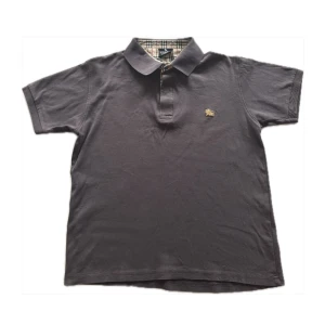 Piké Burberry - Säljer en stilren grå pikétröja från Burberry med klassiskt rutigt mönster på insidan av kragen. Tröjan är kortärmad och tillverkad i 100% bomull, vilket gör den både bekväm och andningsbar. Perfekt för en avslappnad men ändå elegant look.