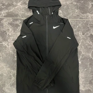 Nike Miller  - Snygg svart vindjacka från Nike med huva och dragkedja. Jackan har långa ärmar och en stilren design med Nike-logga på bröstet. Perfekt för blåsiga dagar den här också håll under armarna man märker inte vet inte hur den fick det faktiskt men den enda defekten 