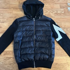 Svart pufferjacka med huva - Snygg svart pufferjacka från Moncler med huva och dragkedja framtill. Jackan har långa ärmar med ett vitt mönster på ena ärmen och ribbade muddar. Perfekt för alla väder