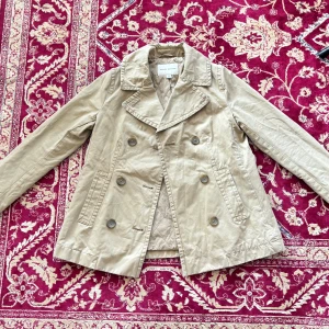 Beige trenchcoat från Banana Republic - Säljer en stilren beige trenchcoat från Banana Republic. Jackan har en klassisk dubbelknäppt design med stora knappar och två fickor framtill. Perfekt för vår och höst! Otroligt fint skick! Kort jacka.