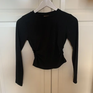 Svart långärmad topp med rynkdetalj - Snygg svart långärmad topp med unik rynkdetalj i midjan som ger en cool look. Perfekt för att styla med jeans eller kjol för en trendig outfit. Materialet är mjukt och bekvämt.