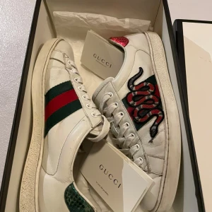 Vita sneakers med ormmönster från Gucci - Snygga vita sneakers från Gucci med ett coolt ormmönster på sidan. Skorna har de klassiska gröna och röda ränderna och är tillverkade i skinn. Perfekta för att ge din outfit en lyxig touch.