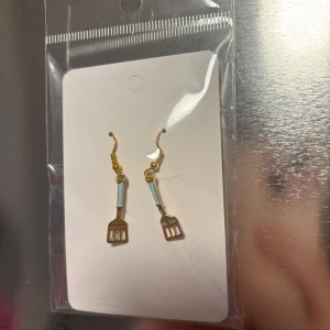 Guldörhängen med stekspade-design - Unika örhängen i guld med en charmig design av små stekspadar. Perfekta för den som vill ha något annorlunda och roligt i smyckesamlingen. De har en liten detalj i blått som ger en extra touch. (Billigt material) 