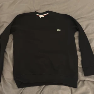 Svart sweatshirt från Lacoste - Säljer en stilren svart sweatshirt från Lacoste med det klassiska krokodilmärket på bröstet. Tröjan har långa ärmar och rund halsringning. Perfekt för en avslappnad stil.