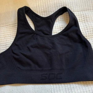 Svart sporttopp från SOC - Säljer en svart sporttopp från SOC. Toppen har en tight passform och är tillverkad i ett syntetmaterial som andas. Perfekt för träning med sin bekväma design och stödjande resår under bysten.