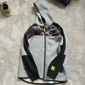 Grå och svart hoodie från Zara Boys - Säljer en cool grå och svart hoodie från Zara Boys med texten 'FUTURE' på ryggen. Den har en dragkedja med en gul detalj och ett camouflagemönster på insidan av huvan. Perfekt för en avslappnad stil.