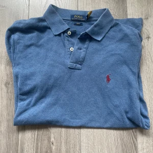 Blå pikétröja från Ralph Lauren - Säljer en klassisk blå pikétröja från Ralph Lauren med den ikoniska loggan broderad i rött på bröstet. Tröjan har korta ärmar och en krage med knappar. Perfekt för en stilren och avslappnad look. Riktigt snygg tröja inför sommar och vår.  Storlek M. Hör av dig vid fundering 🤔 