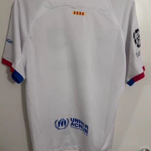 23-24 Vit FC Barcelona fotbollströja - Säljer en vit FC Barcelona fotbollströja från Nike med klubbens emblem och sponsorloggor. Tröjan har korta ärmar med röda och blå detaljer. Perfekt för fans som vill visa sitt stöd.