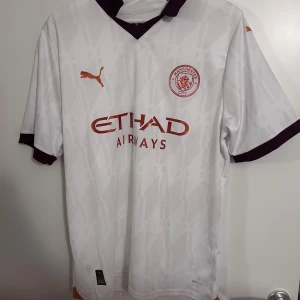 Manchester City fotbollströja - Säljer en vit fotbollströja från Puma med korta ärmar. Tröjan har ett diskret mönster och detaljer i rött, inklusive logotyp och text. Perfekt för fotbollsfans som vill visa sitt stöd.