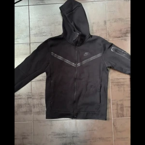 Svart Nike tech hoodie - Säljer en svart nike tech hoodie. Den har en diskret randig detalj över bröstet och en ficka med dragkedja på ärmen. Perfekt för en sportig look! Mycket bra skick utan defekter och sparsamt använd. Storlek M. Pris kan diskuteras 