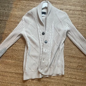 Beige stickad kofta från R-Editon - Säljer en stilren beige stickad kofta från Ralph Lauren med ett snyggt kabelstickat mönster. Koftan har långa ärmar och knappar framtill. Perfekt för sommar och vårkvällar