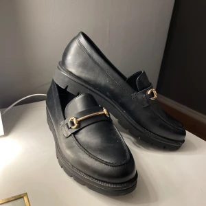 Svarta loafers med gulddetalj - Snygga svarta loafers i skinn med en elegant gulddetalj på ovansidan. Skorna har en robust sula som ger bra grepp och komfort. Perfekta för en stilren look. Använda endast en gång. 