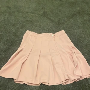 Rosa kjol från Divided - Söt rosa kjol från Divided i en härlig kort modell. Kjolen har en knappdetalj i midjan och är tillverkad i en mjuk blandning av polyester och viskos. Perfekt för en somrig look!