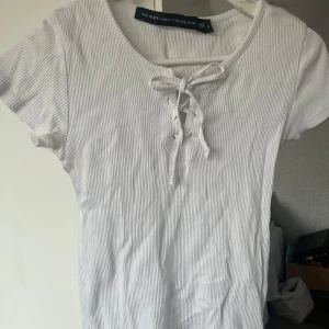 Vit ribbad topp från The Black Label Collection - Säljer en vit ribbad topp från The Black Label Collection. Toppen har korta ärmar och en snygg knytdetalj framtill. Perfekt för en stilren och avslappnad look.