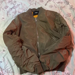 Olivgrön bomberjacka från BikBok - Snygg olivgrön bomberjacka från BikBok med dragkedja framtill och en cool orange insida. Jackan har långa ärmar och ribbade muddar vid ärmslut och nederkant. Perfekt för en avslappnad stil.