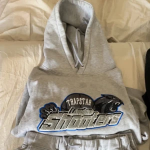 Grå hoodie från Trapstar - Säljer en grå hoodie från Trapstar med trycket 'London Shooters' i svart och blått. Hoodien har en klassisk passform med en stor ficka framtill och justerbar huva. Perfekt för en avslappnad stil.