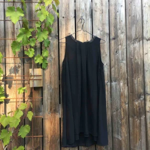  - Oanvänd. Löst sittande svart klänning med halsringning dekorerad med pärlor. Dekorativ vikning mitt fram. Slutar strax ovanför knäna. En aning längre bak. Skirt övre tyg och skön ”underklänning”.  Kan stylas med skärp i midjan.