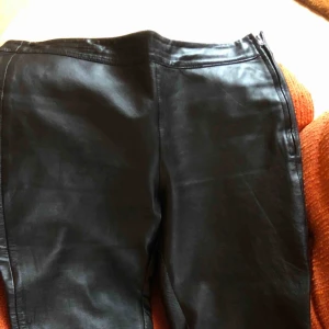  - Svarta skinnbyxor. Ej märkta men passar en storlek 40. Knäpps i vänster sida med dragkedja och tryckknapp. Inga fickor eller hällor. 