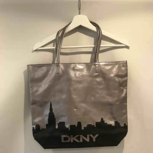 Silvrig DKNY väska - Supersnygg silvrig väska med svart stadssilhuett på från DKNY! Säljer eftersom den får alldeles för lite uppmärksamhet i min garderob.