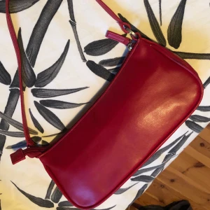 Liten axelremsväska från Åhléns  - Liten röd axelremsväska i trendig modell. Från Åhléns. I gott begagnat skick. Köparen står för frakten. 