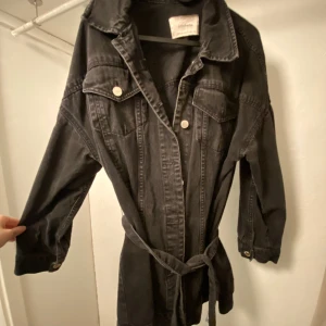 Stradivarius jeansjacka - Jeansjacka i färgen svart/grått med fickor, skärp o knappar. Liten defekt, Där man lägger skärpen, ena sidan har lossnat av men man kan sy ihop den. Kan skicka tydligare bild om önskas