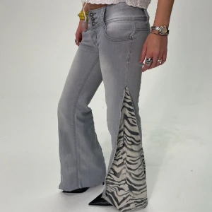 REMAKE ZEBRA JEANS - Storlek: Midja (70cm), Innerbenslängd (82cm), total längd (101cm). Grå, remake, lågmidjade, bootcut jeans med vit/svart zebramönstrad slit. Modellen är 172 cm.