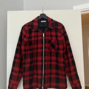 J.Lindeberg  - En snygg och stilren skjorta med rött och svart rutmönster. Den har en dragkedja framtill och en ficka på bröstet. Skjortan är perfekt för både vardag och fest.