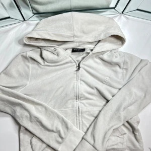 Juciy couture, ROBERTSON GOLD HOODIE - Ziptröja med luva i velour från Juicy Couture Försluts med dragkedja framtill Mudd i ärmslut samt nederkant Två fickor framtill. Kommer utan snören vid luvan.