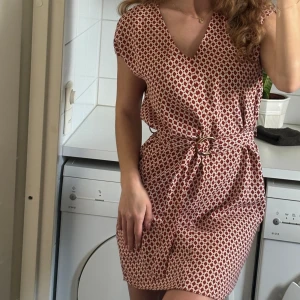 Kortärmad mönstrad klänning med skärp - Nyskick! Använd 1-3 gånger. Mönstrad klänning i lätt material, skärp med guldigt spänne. Vit och lite terrakotta röd färg. Storlek XS/34.