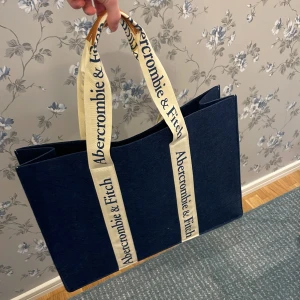 Tygväska, Abercrombie & Fitch - Relativt stor tygväska i ett marinblått mjukt tyg. Handtag med branding och detaljer i konstgjort läder.