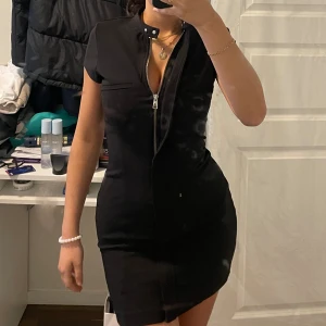 Svart klänning med dragkedja - Säljer denna svarta klänning som har en dragkedja hela vägen och två knappar vid kragen som man kan styra och ställa med. Köpt från Zara för ungefär 6-7 månader sedan. Den är som ny få jag använt den 1-2 gånger. Den har en tunn klänning under.