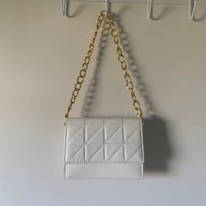 Vit axelväska  - Vit axelväska med sydda detaljer och guldkedja. Själva väskan är 20x17 cm och kedjan är 72 cm. Tyvärr har väskan fått några märken på baksidan se bild 3. Annars en mycket rymlig och snygg väska.🤍köpt från en annan användare här på plick.🤍💛