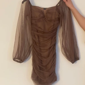 Brun klänning  - Bodyconklänning med veckad mesh och som framhäver fint kroppen. Finns även med volangärmar som ger en extra detalj. Vid bröstet har det ett ”v”. 