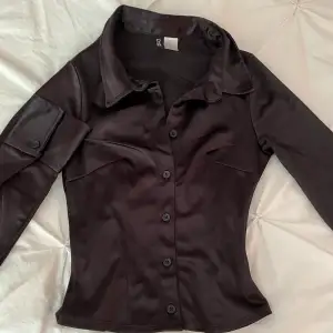 En oanvänd blus/skjorta från H&M i svart/mörkbrun nyans. Satin liknande material, storlek XS. Plagget har en ganska figurnära passform. 