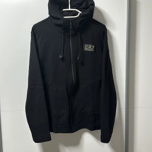 Svart jacka från Emporio Armani - En svart zip hoodie från Emporio Armani med dragkedja och huva. Hoodien har två framfickor. Köpt i butik för 2399kr. Använd 5-6 gånger vid bra tillfällen. Storlek S/M.