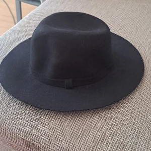 Svart hatt - En elegant svart hatt med bred brätte. Hatten har ett enkelt band runt kullen och är tillverkad i ett mjukt material. oanvänd. 100% ny.