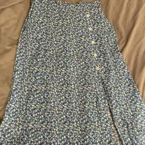 Lång blå kjol med blommönster och och knappar på sidan. Kjolen har en slit på sidan där knapparna sitter🌸 kjolen har djupa fickor🤩 köpt på Lindex och sparsamt använd. Jättefin till våren/sommaren!😍😍