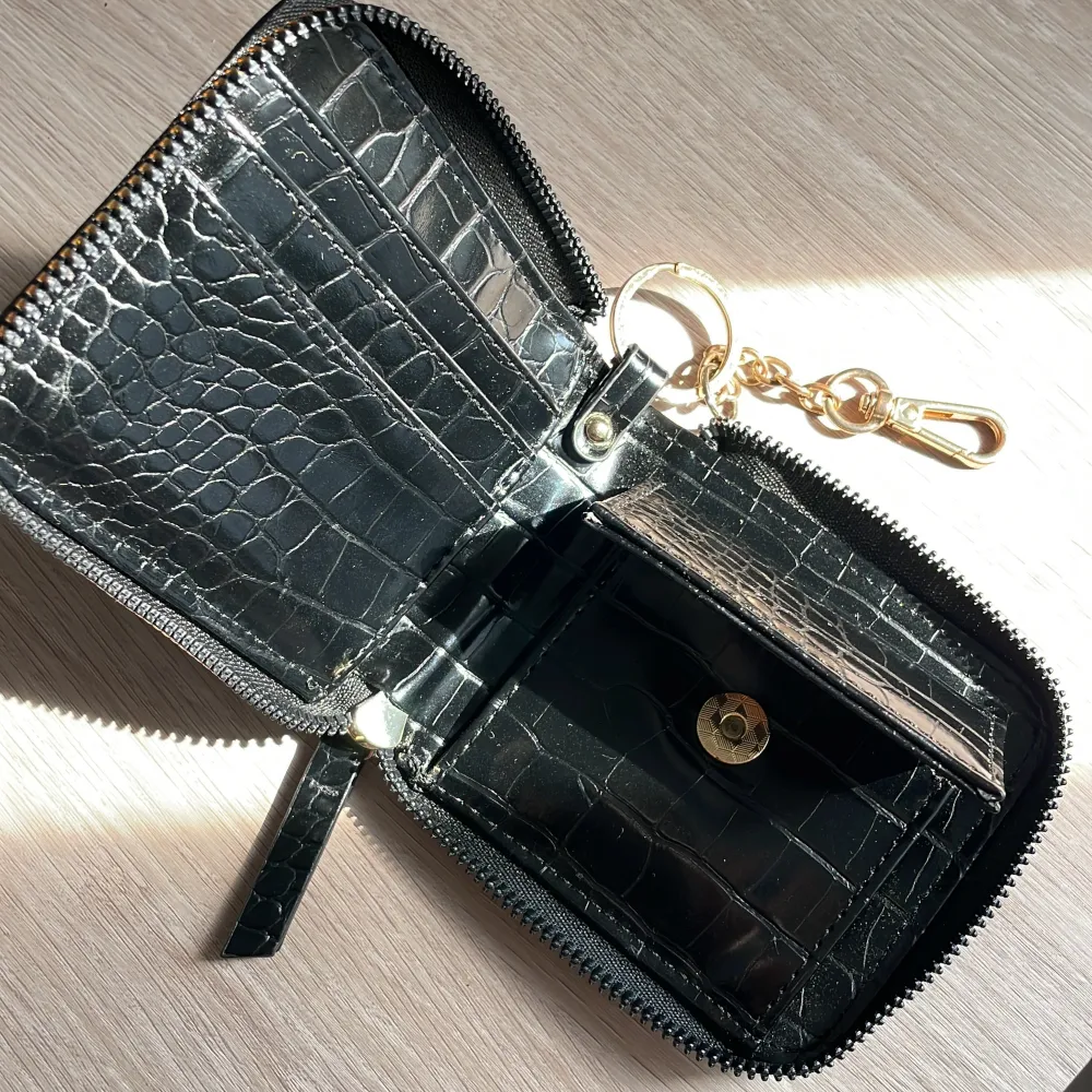 En elegant svart plånbok med krokodilpräglat mönster. Plånboken har en dragkedja runtom och en guldfärgad kedja med karbinhake. Insidan har flera fack för kort och en knappstängd ficka för mynt.. Laukut & Käsilaukut.