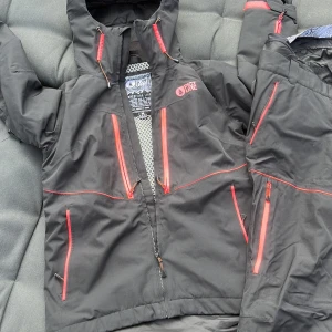Svart jacka och skidbyxor från Picture - En svart jacka från Picture's Expedition Line med rosa dragkedjor och detaljer samt skidbyxor till. Jackan har en huva och flera fickor med dragkedjor. Den är tillverkad av tekniska material för att klara av tuffa väderförhållanden. Jackan är storlek M och byxorna L då de är ganska små i storleken skulle jag säga att de är ca  stl 40.