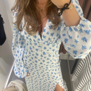 Vit klänning med blå blommor - En elegant vit klänning med blå blommor. Klänningen har långa ärmar och en slits på sidan. Den har knappar framtill och en v-ringad hals.