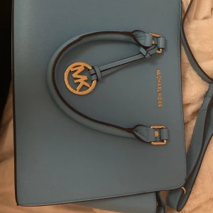 Blå handväska från Michael Kors - En elegant blå handväska från Michael Kors med gulddetaljer. Väskan har en dragkedja och ett rymligt inre fack. Den är tillverkad av skinn och har en klassisk design med Michael Kors logotyp på framsidan.