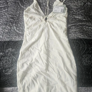 Vit klänning från H&M - En elegant vit klänning från H&M med smala axelband och en liten öppning framtill. Klänningen är tillverkad av 98% återvunnen polyester och 2% elastan, med en foder av 50% återvunnen viskos.