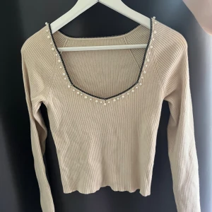 Beige ribbad tröja med pärldetaljer - En beige ribbad tröja med långa ärmar och pärldetaljer runt halsringningen. Tröjan har en v-ringad design och är tillverkad i ett stickat material.